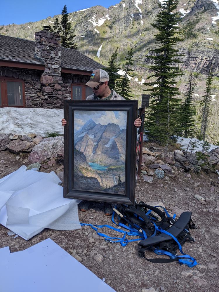 A man in a ball cap holding a large framed painting outside a stone and timber building with alpine trees and mountain ridges in the background.