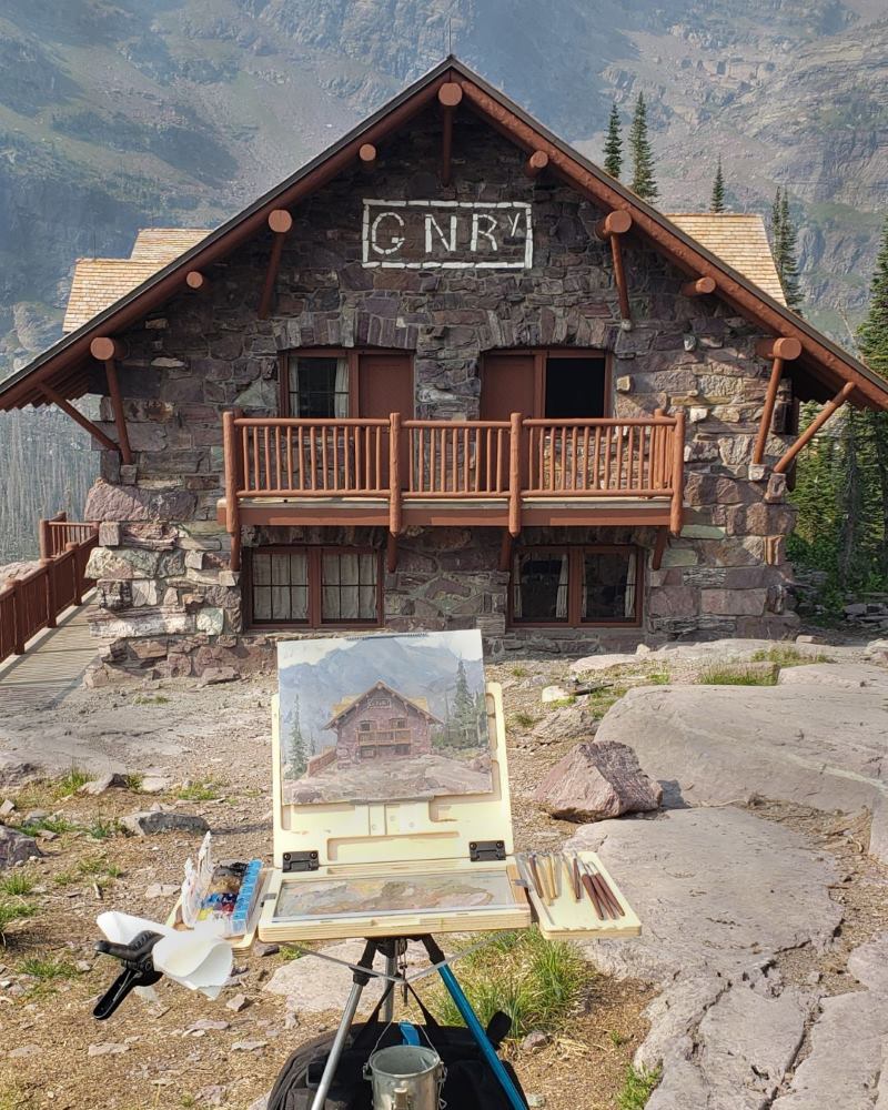 A rock brick and timber building with GNR spelled out in white stone at the top of the eave with a small easel and a plein air painting in the foreground.