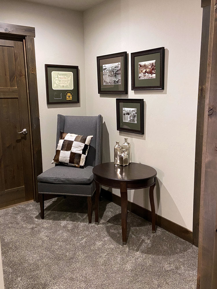 A sitting area in a home with a grey chair and side table. The doors and trim are wooden and the walls are painted white. Three vintage photographs and one certificate and patch are all framed cohesively and hung on the wall above the chair.