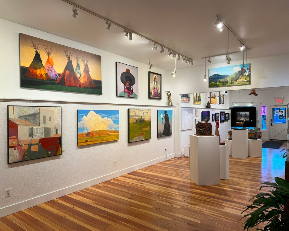 A view of the front room of FoR Fine Art gallery in Whitefish. Large, colorful, contemporary western art is hung along a white wall with a few pedestals featuring sculptures. The room has green potted plants and natural wood floors.
