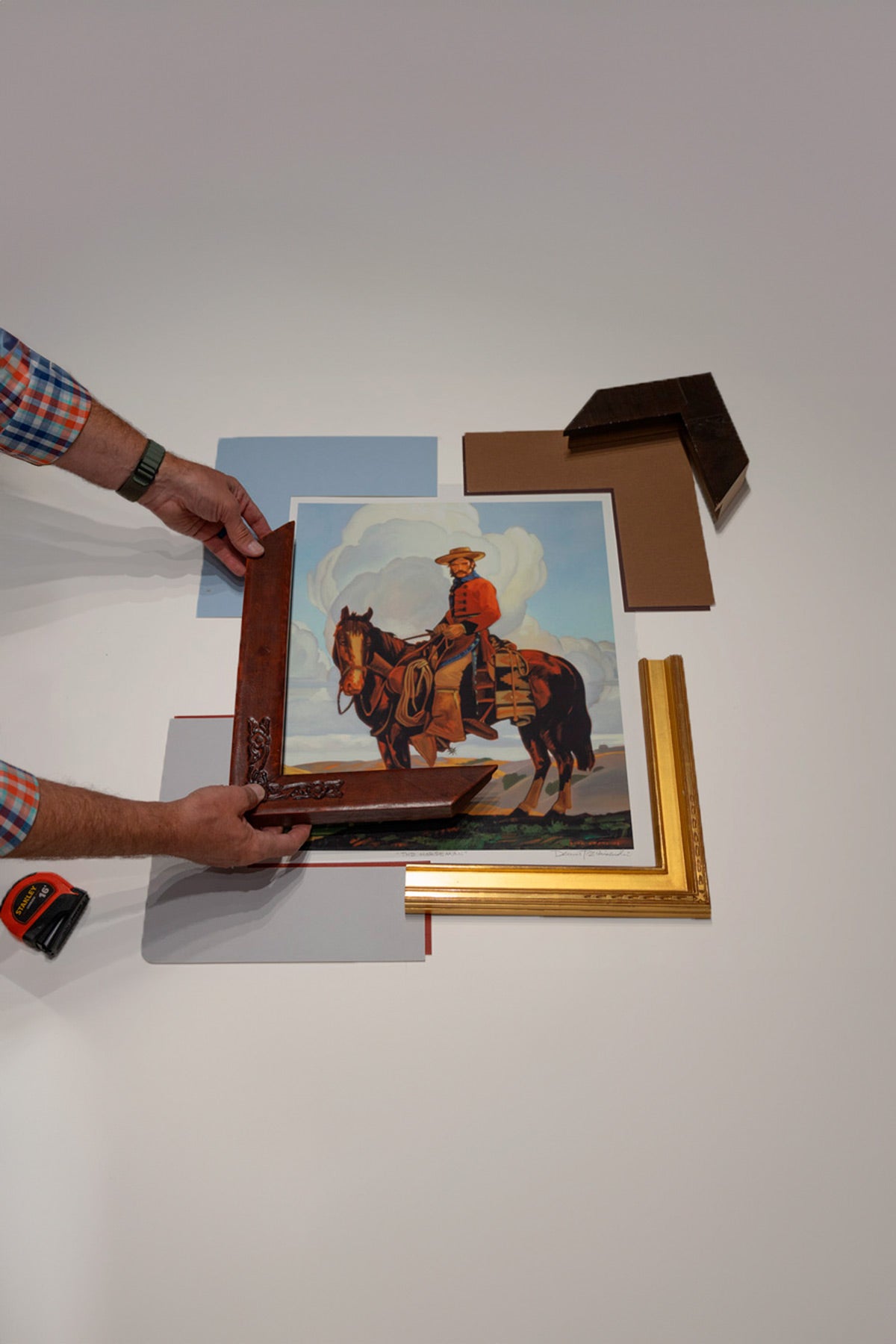 Two hands holding a corner sample of leather moulding next to a western print of a cowboy on a horse. The print is on a white table with various mats and moulding samples around it.