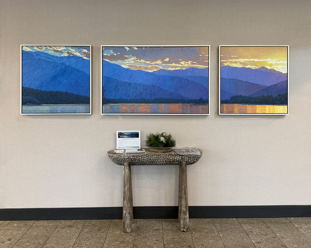 A triptych of oil paintings featuring mountains above a lake during a golden sunset hung above a small table made of wood in a hotel lobby.
