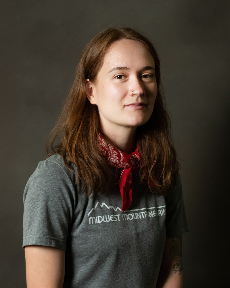 A woman with red hair wearing a grey t-shirt and red bandana softly smiling.