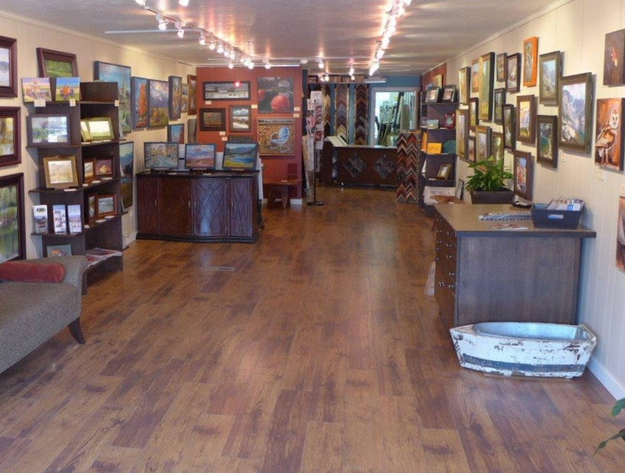 A small gallery and frame shop filled with small- to medium-sized artwork, a framing design area with moulding and mat samples, and a few pieces of dark wood furniture for displays.