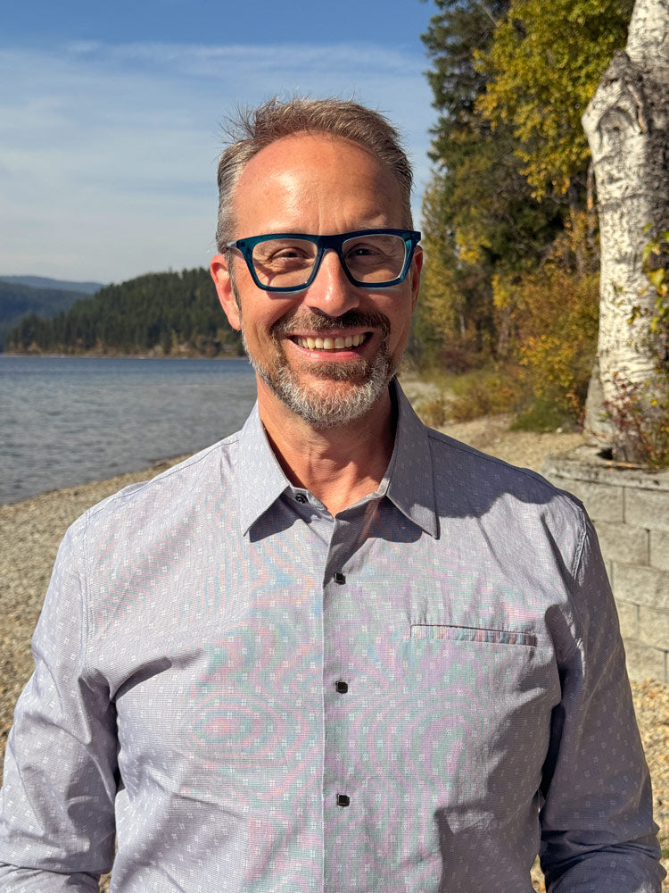 A smiling man with short grey hair, a trimmed beard, and bold blue glasses.