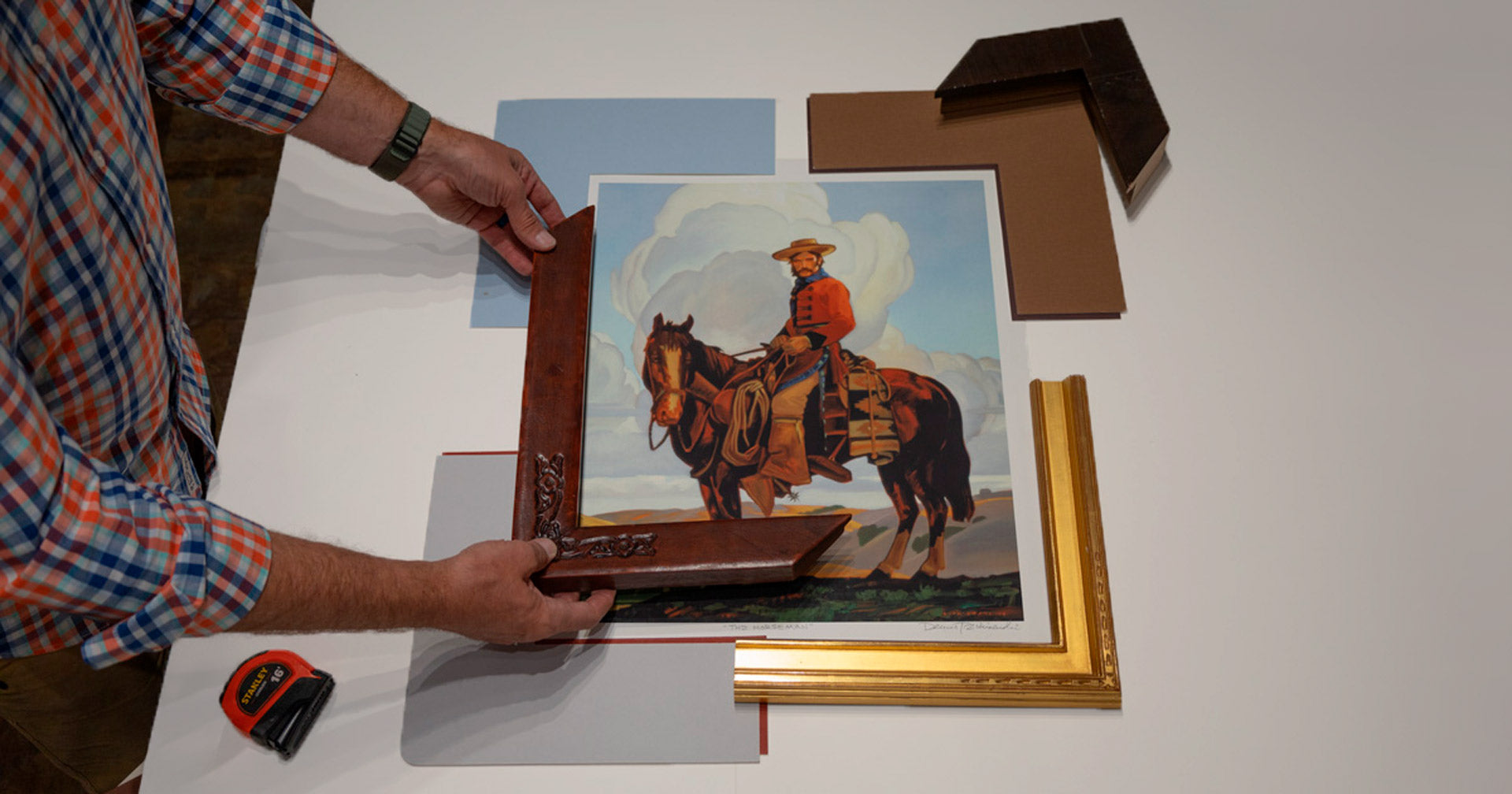 Two hands holding a corner sample of leather moulding next to a western print of a cowboy on a horse. The print is on a white table with various mats and moulding samples around it.