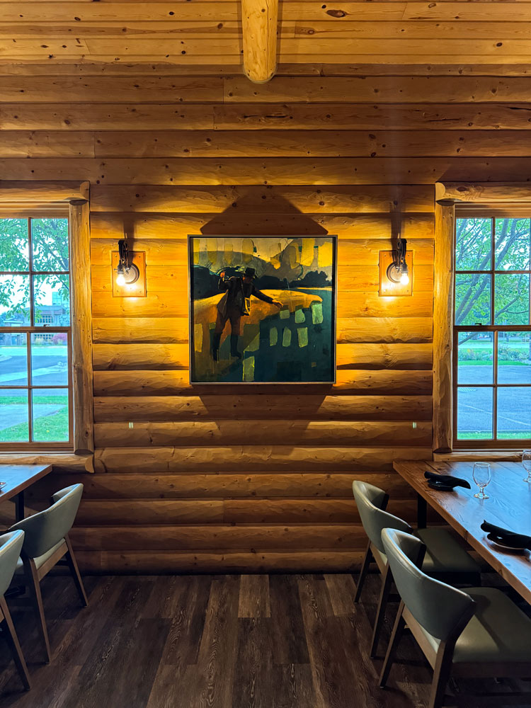 A square mixed media piece depicting a man casting a fly rod. The painting is hung on a log wall above a table in a restaurant with two lights on either side.