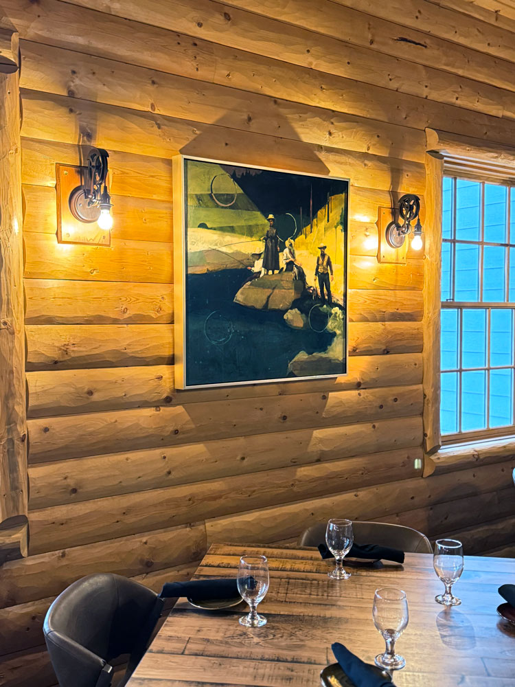 A square mixed media piece depicting a vintage image of a family standing on rocks fishing. The painting is hung on a log wall above a table in a restaurant.