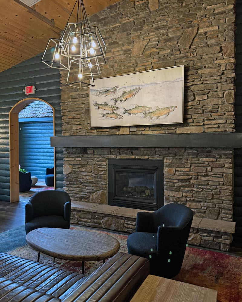 A view of a large stone fireplace with deep blue and brown furniture, a geometric chandelier, and a large ink wash painting above the mantel depicting six bull trout swimming across the canvas.