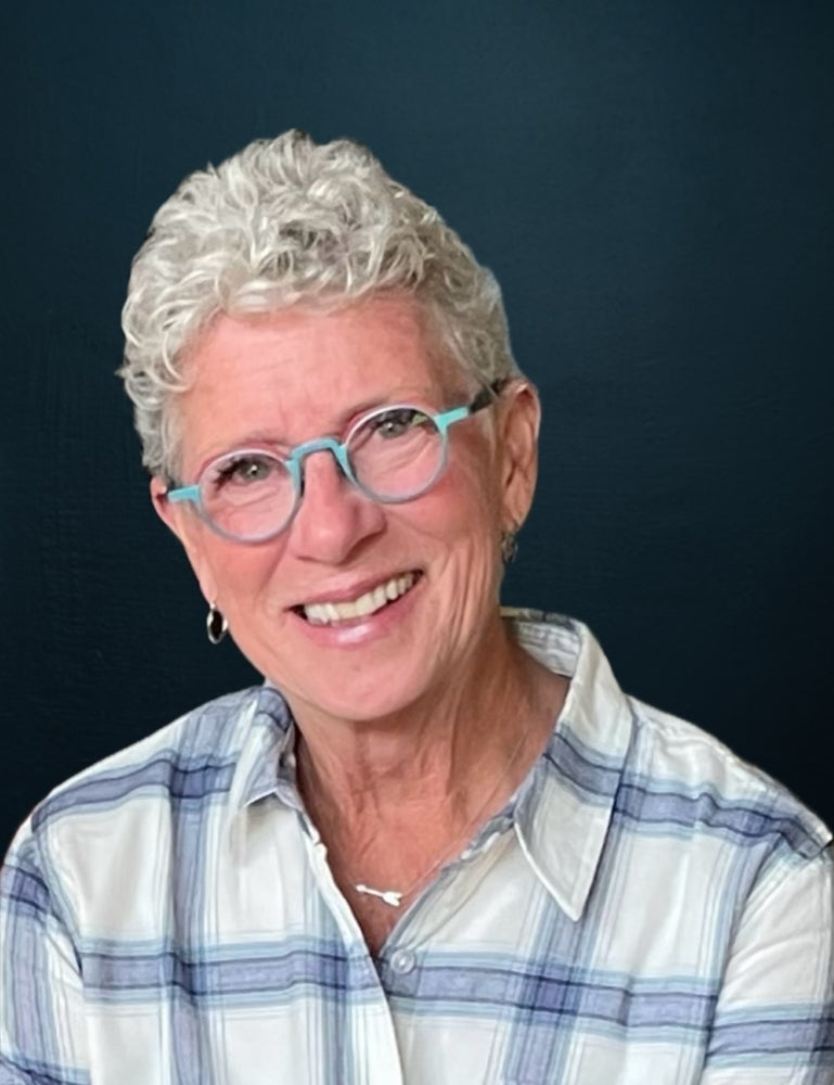 A headshot of a woman with short white hair and round, teal glasses frames. She is wearing a plaid shirt and smiling.