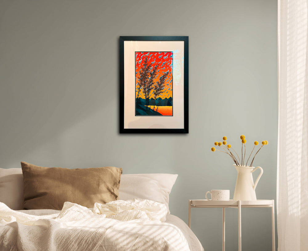 A framed watercolor with a bold orange and yellow sunset, mountains, and silhouetted trees in the foreground is hung above a bed and nightstand with light, neutral colors.