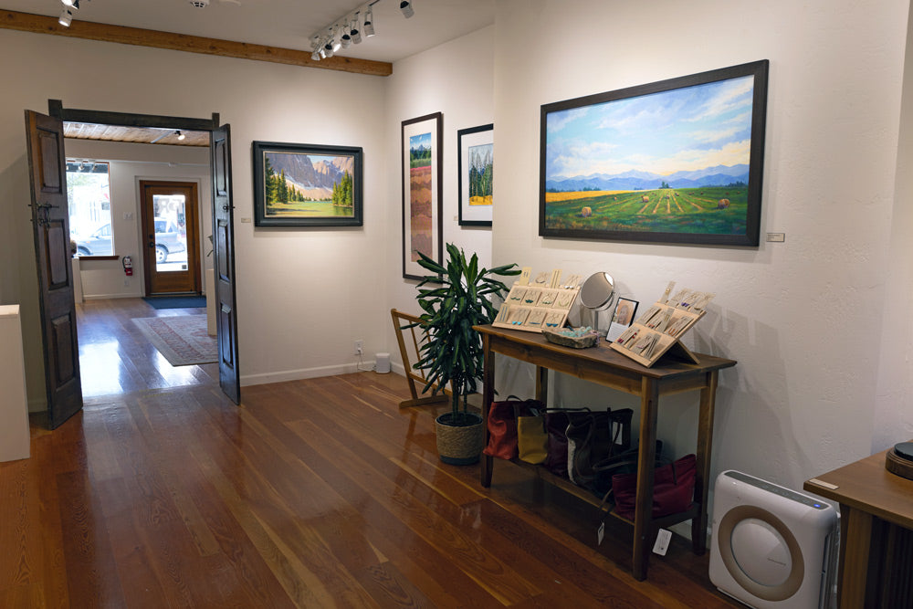 A warm white gallery wall with colorful paintings hung above a row of small bronze sculptures, wooden bowls, and blown glass.