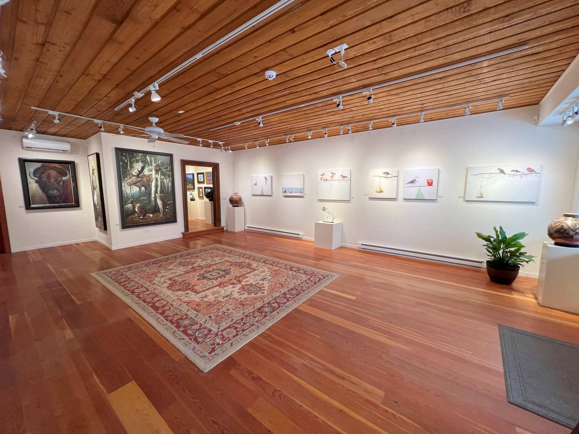 A bright gallery room with wooden floors and ceilings, and area rug, and a few plants. Various pieces of artwork are hung along the walls and a few pedestals hold Mata Ortiz pottery and contemporary sculptures.