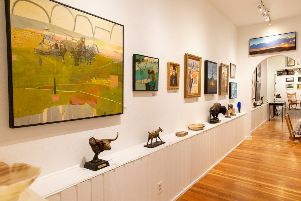 A warm white gallery wall with colorful paintings hung above a row of small bronze sculptures, wooden bowls, and blown glass.
