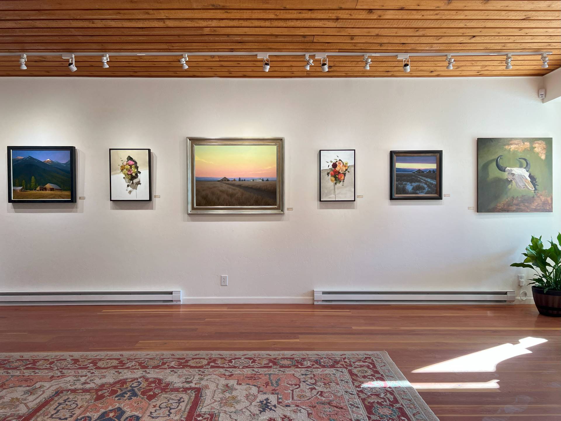 A warm white gallery wall with wood panel ceiling and floor and a large area rug. The wall is hung with large artworks by Richie Carter ranging from moody Montana landscapes, contemporary florals, and realism still life.