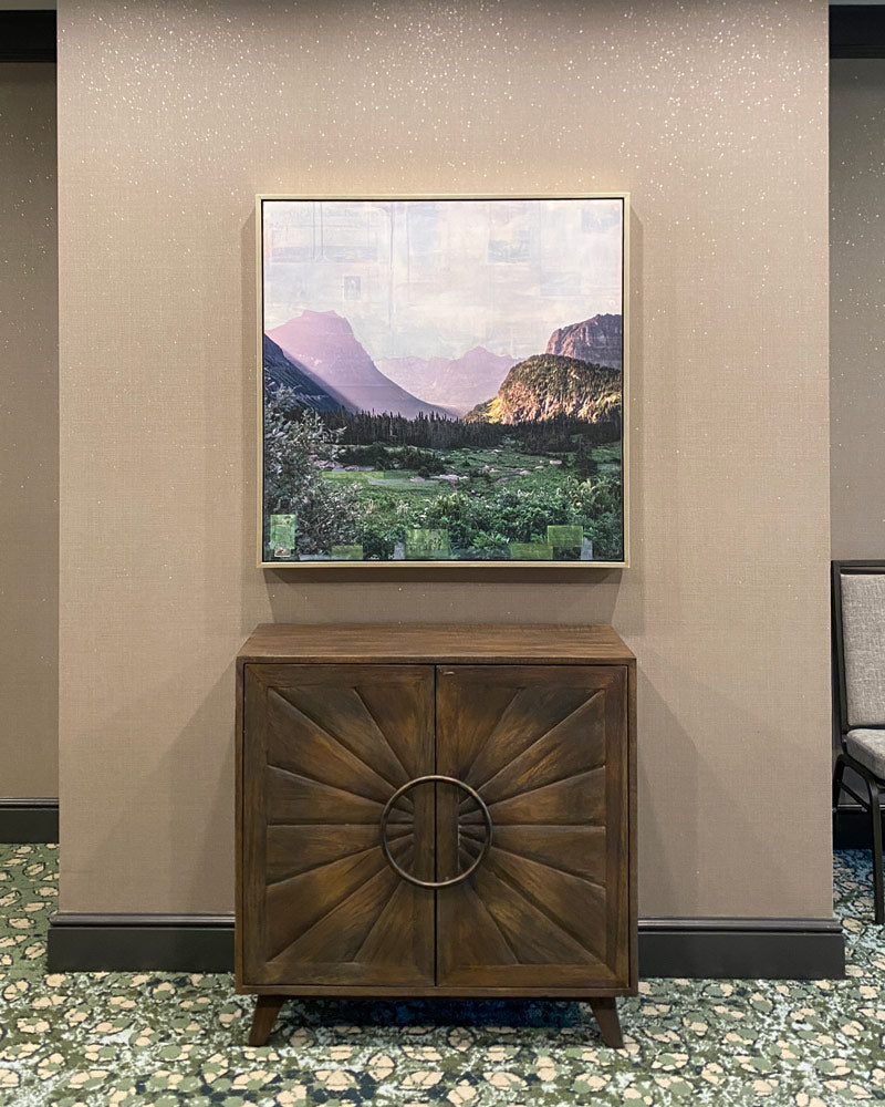 A mixed media piece with soft purple mountains fading to the horizon and rich green foliage in the foreground. The artwork is hung above a dark brown end table.