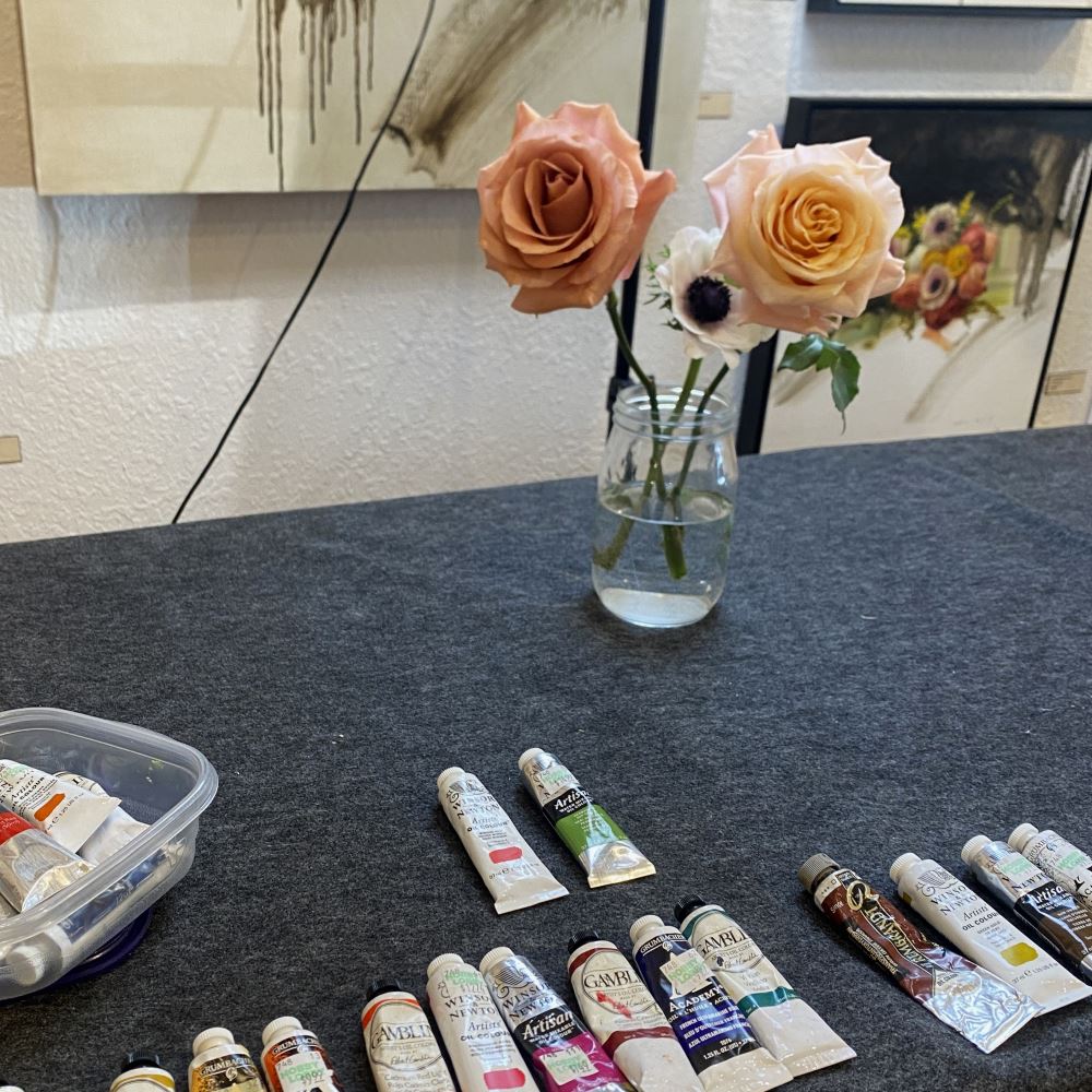 A table with a grey cloth on top of it and a row of small paint tubes of different colors. The far edge of the table has a mason jar with water and three flowers.
