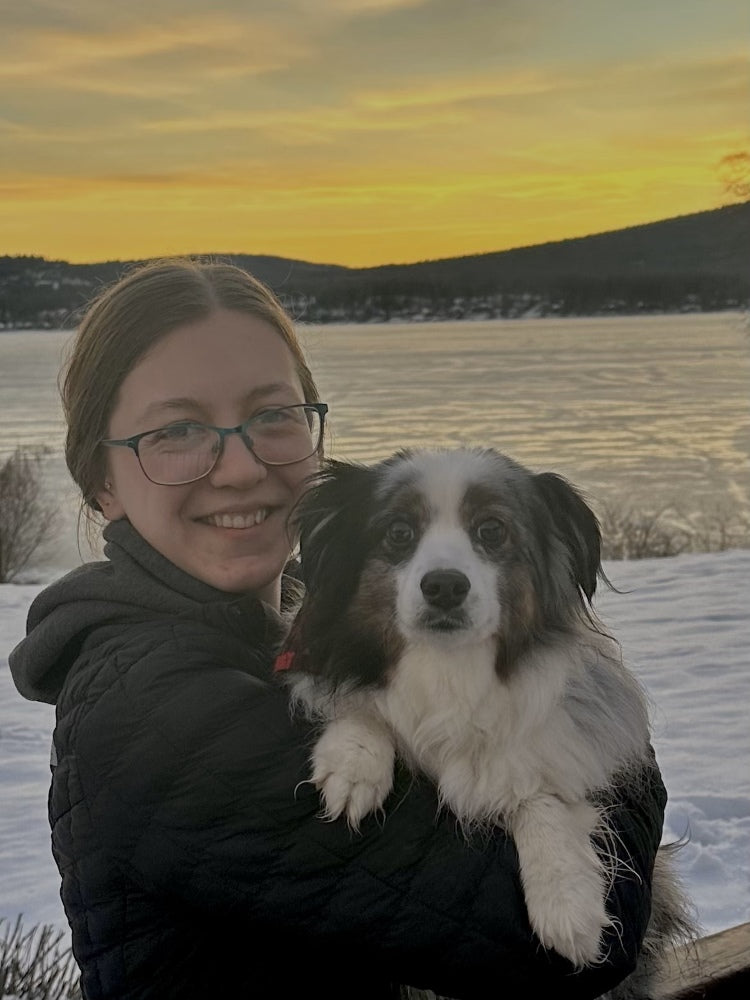 A woman wearing a cap, sunglasses, and a cream long sleeved shirt holds a colorful Rice Krispie treat with a pink candle on top of a mountain.
