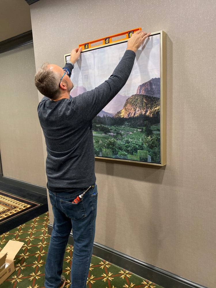 A man holds a level on the top of a JC Spock landscape hung on a beige wall in the Hilton Garden Inn.