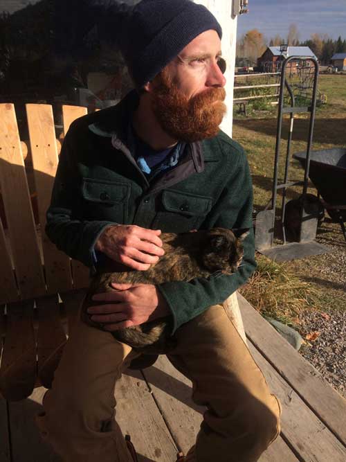 A man with a red beard, blue beanie, and green coat sitting outside on a bench while holding a tortoiseshell cat.