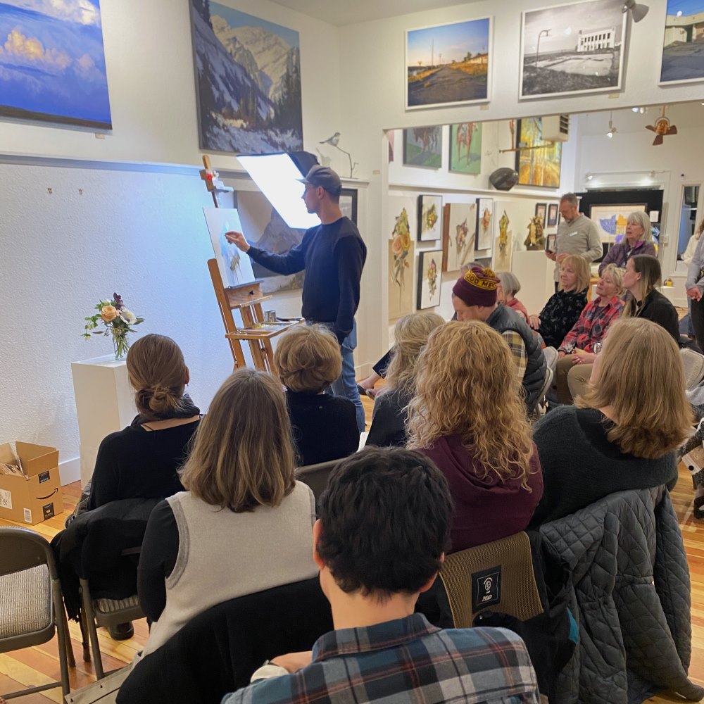 a group of people watch Richie carter being a contemporary floral painting demo on an easel with a floral arrangement on a pedestal next to the artist. The group is in a gallery with white walls and colorful artwork.